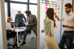 Two side by side images of a workplace. On the left side a group of people are having a meeting at a table. On the right side, two people are planning/writing information on a white board.