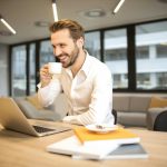 Man drinking a hot drink smiling at his desk.